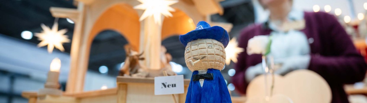Im Vordergrund ist das geschnitzte Leipziger Messemännchen zu sehen, im Hintergrund sind – leicht verblasst – weitere Holzfiguren sowie die Ausstellerin auf der HANDWERK live zu erkennen.