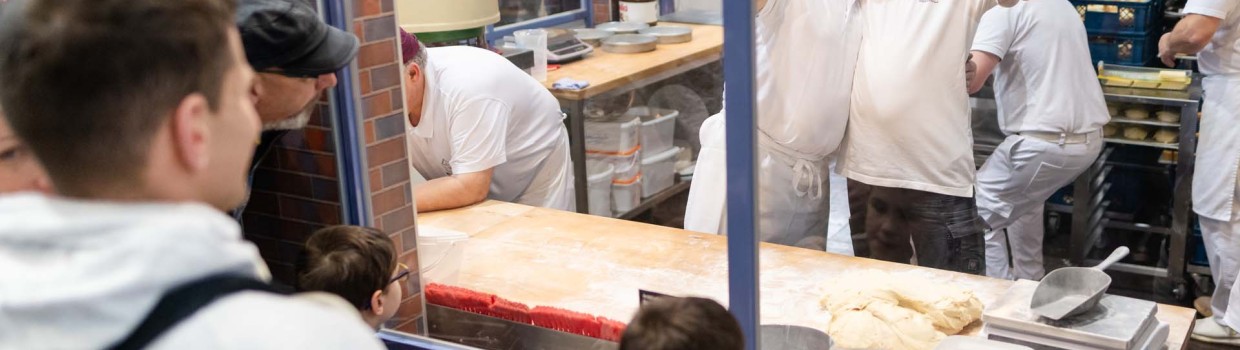 Mit lustigen Grimassen winken zwei Bäcker hinter einer Glasscheibe in die Kamera, während Besuchende der HANDWERK live die Gläserne Bäckerei bestaunen.