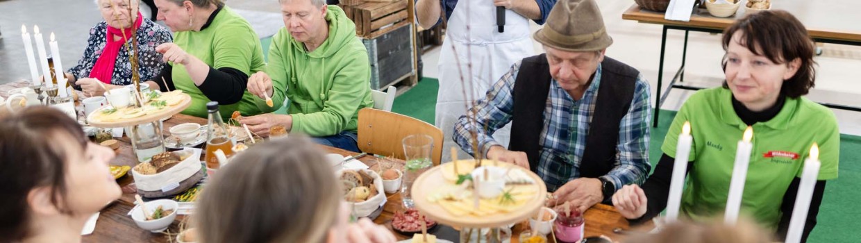 Mehrere Besuchende der HAUS-GARTEN-FREIZEITsitzen an einem gedeckten Tisch, essen gemeinsam und unterhalten sich, während im Hintergrund ein Aussteller mit Mikrofon erklärt.