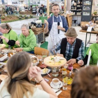 Mehrere Besuchende der HAUS-GARTEN-FREIZEITsitzen an einem gedeckten Tisch, essen gemeinsam und unterhalten sich, während im Hintergrund ein Aussteller mit Mikrofon erklärt.