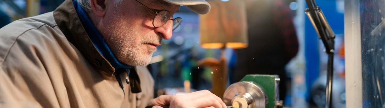 Ein Mann mit Brille und Cap drechselt auf der HANDWERK live an einem beleuchteten Tisch detailliert an einem Holzstück.