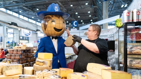 Auf dem Bild zeigt ein Aussteller der HAUS-GARTEN-FREIZEIT dem Leipziger Messemännchen die verschiedenen Käsesorten seines Standes, und das Messemännchen duftet daran.