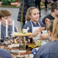 An einem gedeckten Tisch auf der HAUS-GARTEN-FREIZEIT sitzen mehrere Besuchende im Gespräch beisammen, während auch ein Junge und ein Mädchen mit am Tisch zu sehen sind.