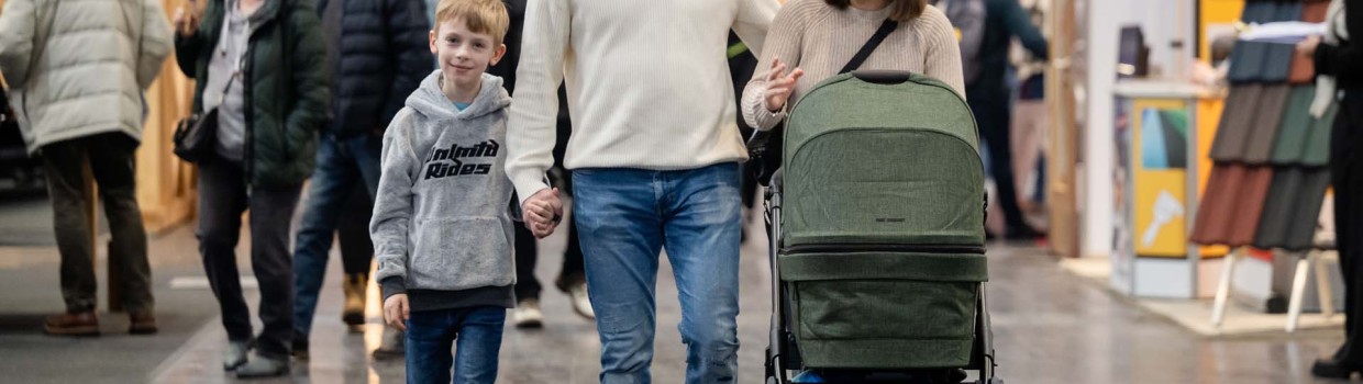 Zu sehen ist eine Familie (Eltern, ein kleiner Sohn und ein Kinderwagen), die über die HAUS-GARTEN-FREIZEIT geht.