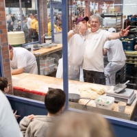 Two bakers behind a glass pane wave at the camera with funny faces, while visitors to HANDWERK live marvel at the glass bakery.